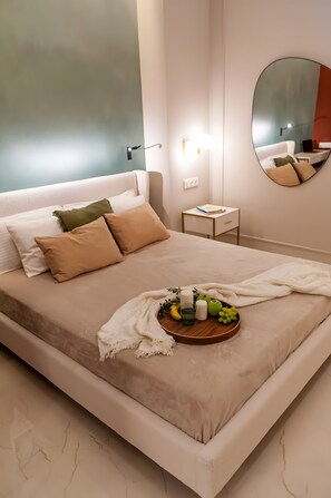 A neatly made bed with a tray of fruits and a candle, a bedside table with a lamp, and a large mirror on the wall.