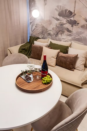 A round dining table with a white tablecloth, a bottle of wine, two glasses, a wooden tray with grapes and chocolate, and a green plant.