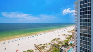 Blick auf einen Strand von einem Hochhaushotel, mit blauem Himmel, sandigem Strand mit Sonnenschirmen und einem Poolbereich.