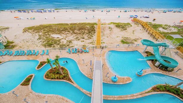 Un parque acuático con toboganes y piscinas múltiples, una zona de playa y un puente de madera que conecta las piscinas con la playa.