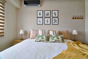 A neatly arranged bedroom with a bed, bedside lamps, a wall-mounted air conditioner, and framed botanical prints.