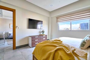 A modern bedroom with a bed, a dresser, a TV, and a window with a city view.