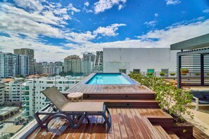 A rooftop pool with a wooden deck, seating, and potted plants.