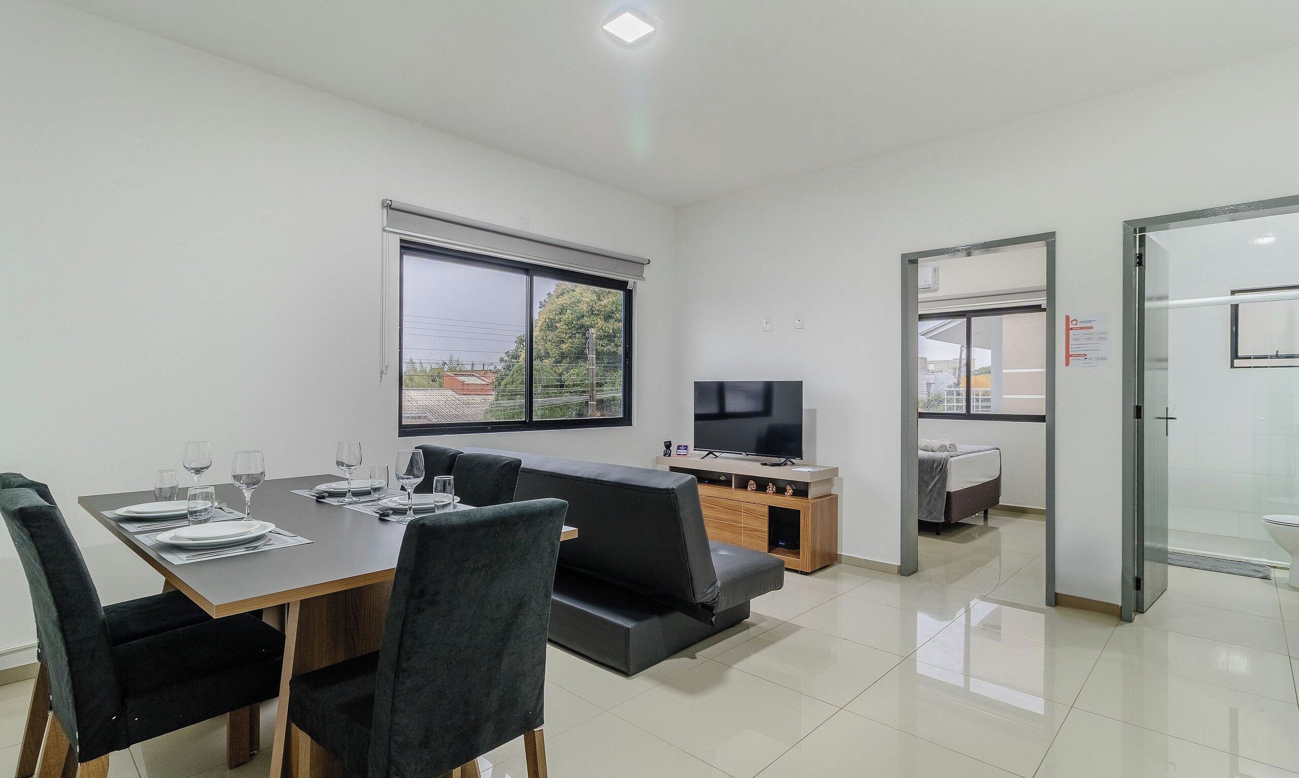 A modern, well-lit room with a dining table set for four, a sofa, a TV, and a separate bathroom area.