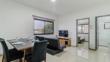 Una habitación moderna y bien iluminada con una mesa de comedor preparada para cuatro personas, un sofá, un televisor y un baño separado.