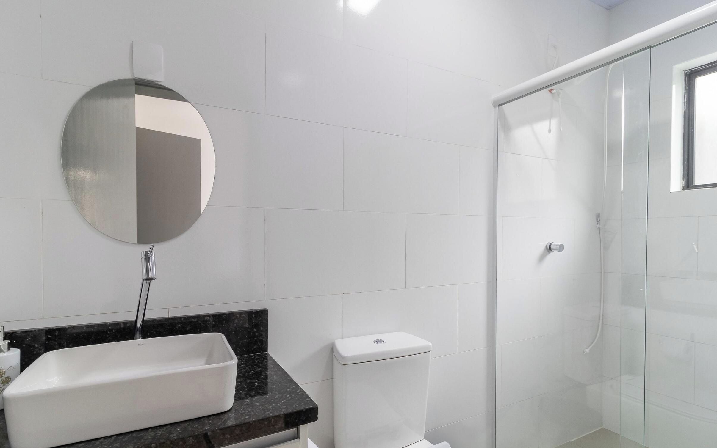 Et badeværelse med hvide fliser, et rundt spejl, en vask og et toilet.