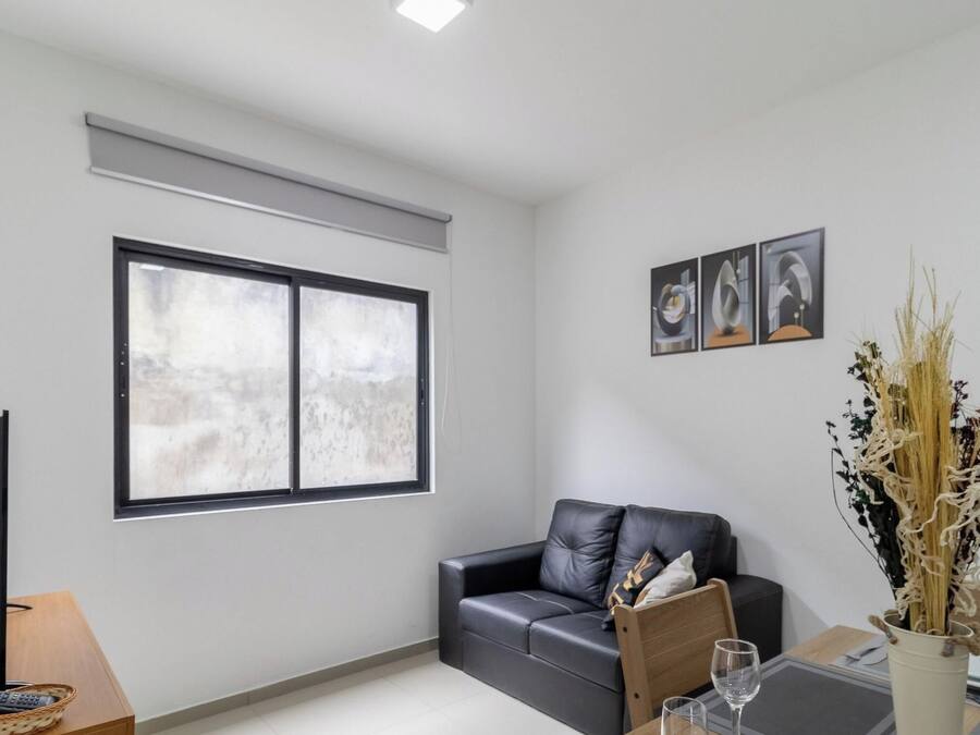 A modern living room with a black leather sofa, a flat-screen TV, a dining table set with plates and glasses, and a large window with a view.
