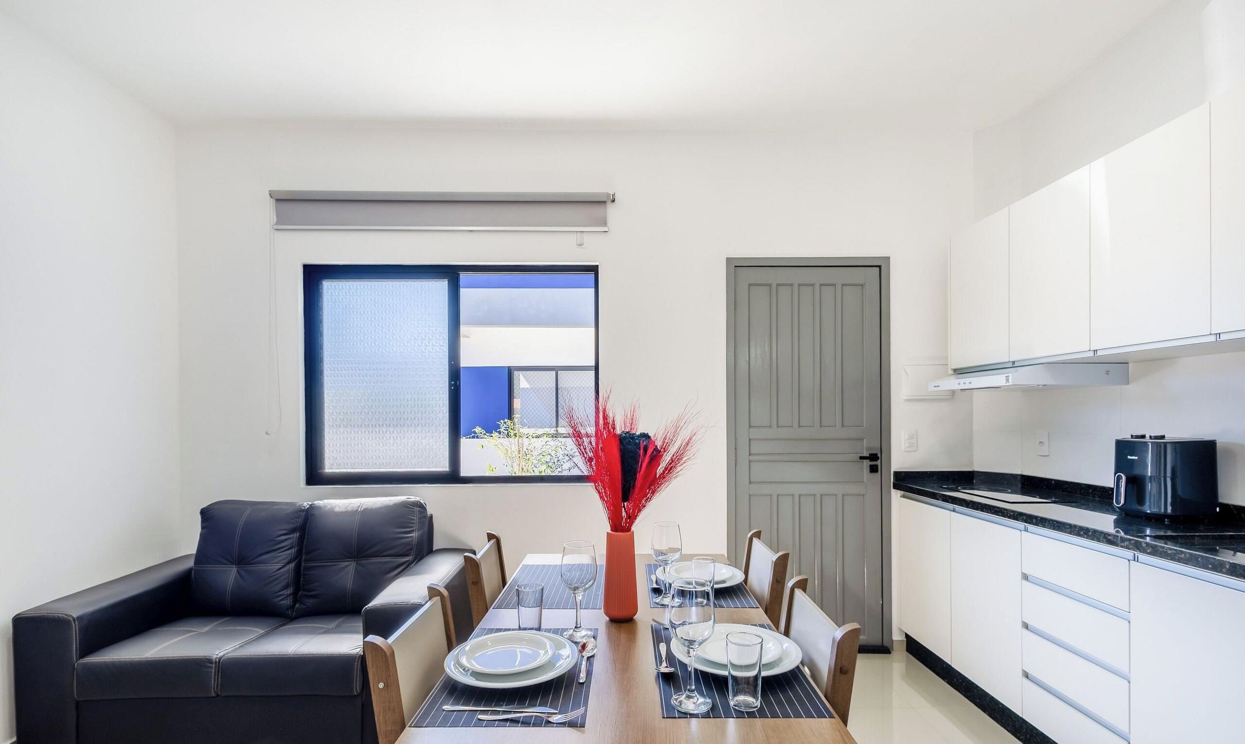 A compact living space with a dining table set for two, a sofa, a kitchen area, and a window with blinds.