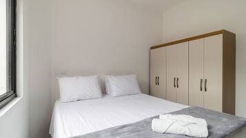 Un dormitorio con una cama, almohadas, una manta gris, una toalla blanca y un armario.