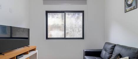 A living room with a flat-screen TV on a wooden stand, a black leather sofa with a cushion, and a window with a view.