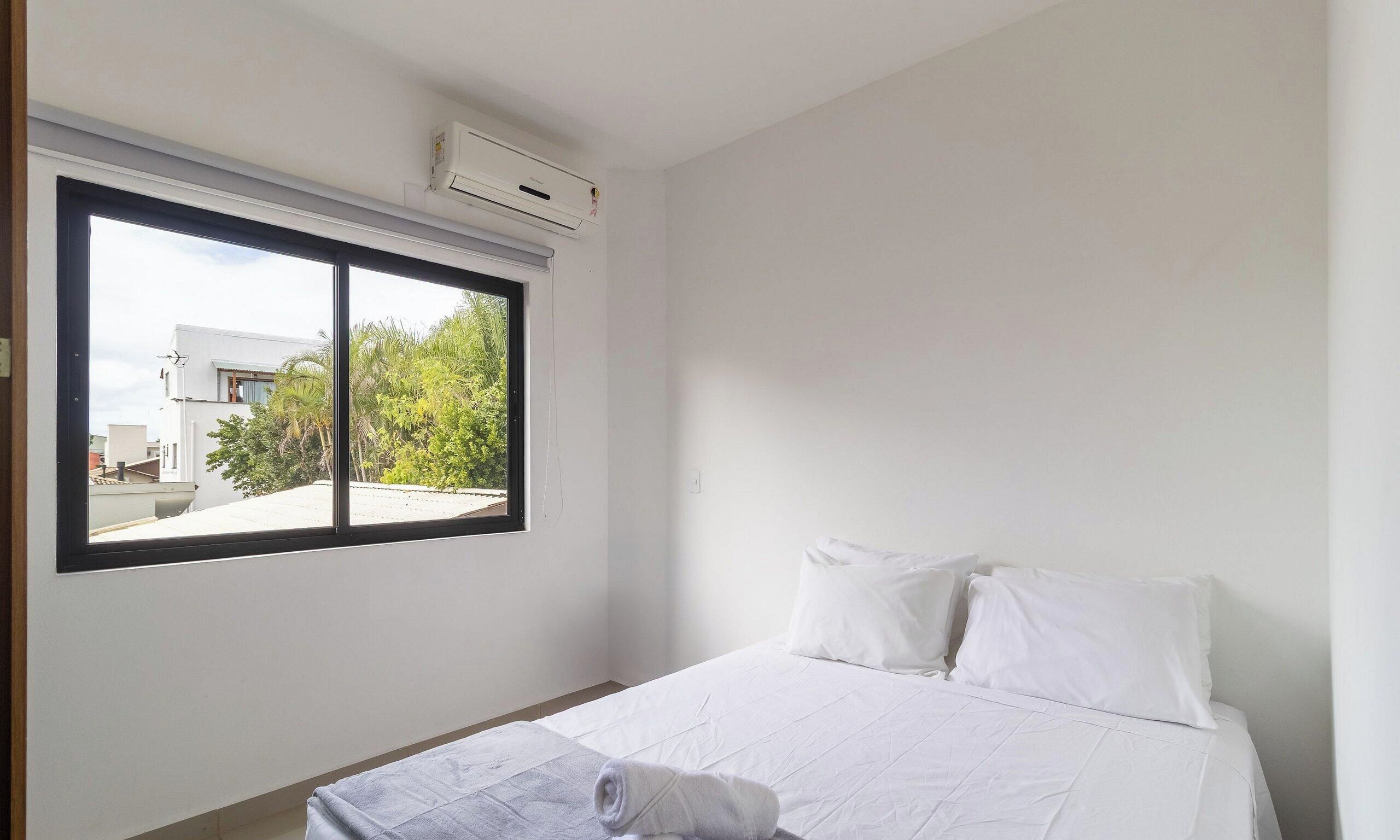 A hotel room with a bed, a window with a view of buildings and trees, and an air conditioning unit on the wall.