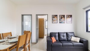 A modern living room with a black leather sofa, wooden dining table with chairs, and a hallway leading to a bathroom. - Beautiful Apartments in Campeche - Floripa - SCO (Florianópolis)