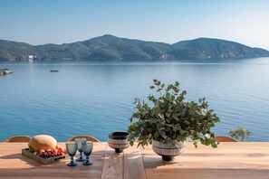 Uma mesa com uma cesta de frutas, copos e um vaso de planta, à beira de um lago com montanhas ao fundo.