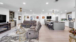 A spacious living room with a large gray sectional sofa, a glass coffee table, and a TV. The kitchen area is visible with white cabinets and a central island.