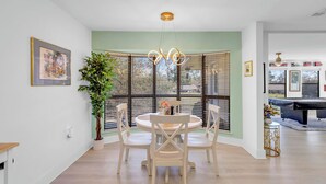 A dining area with a round table and chairs, a chandelier, and a large window with blinds.