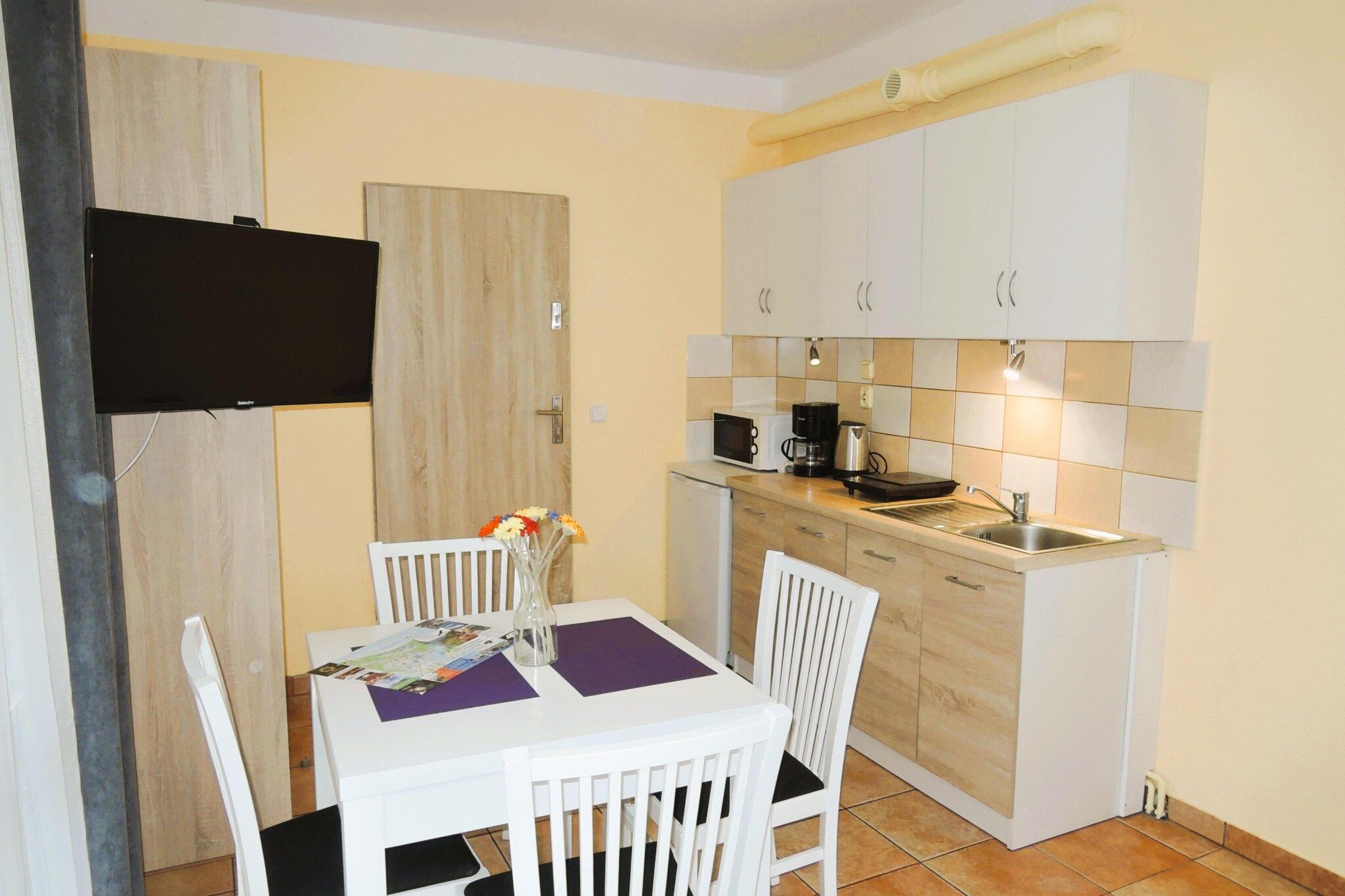 Une cuisine compacte avec un coin repas, comprenant une table avec des chaises, un téléviseur à écran plat fixé au mur et un four à micro-ondes.