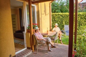 A patio with two people seated at a table, a chair, and a small table with a bottle.