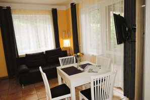 A room with a white table, chairs, a sofa, a TV, and a window with curtains.