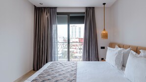 Una habitación de hotel moderna con una cama grande, cortinas grises, vistas de edificios a través de un amplio ventanal y una lámpara colgante.