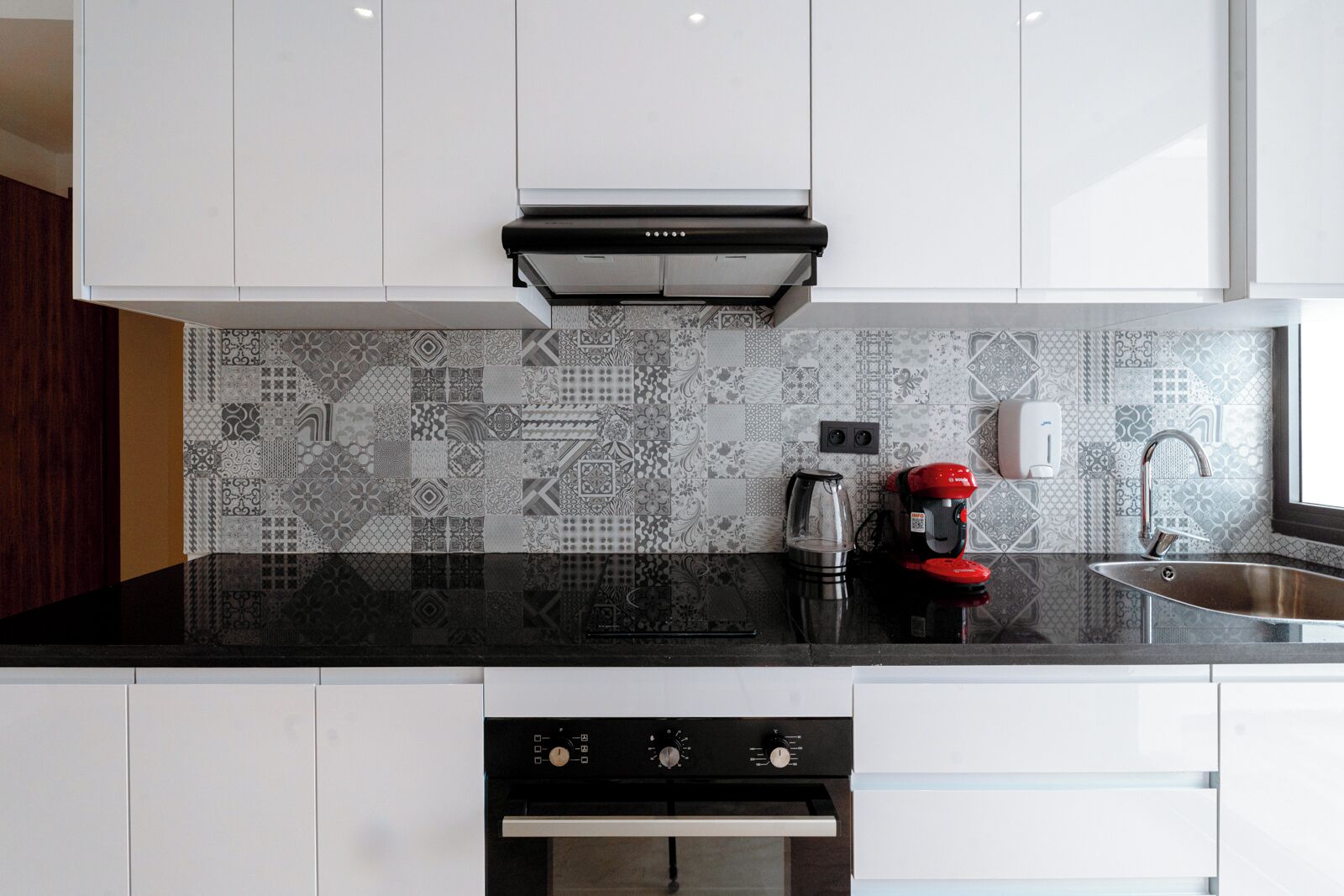 Una cocina moderna con armarios blancos, encimera negra, salpicadero estampado, cafetera y pileta de acero inoxidable.