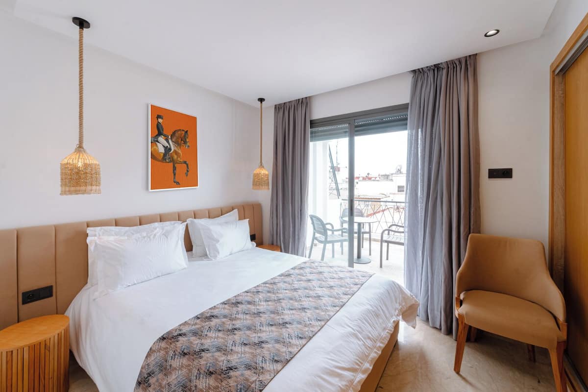 Une chambre d’hôtel avec un grand lit, une chaise, un balcon avec une table et des chaises, et un tableau au mur.