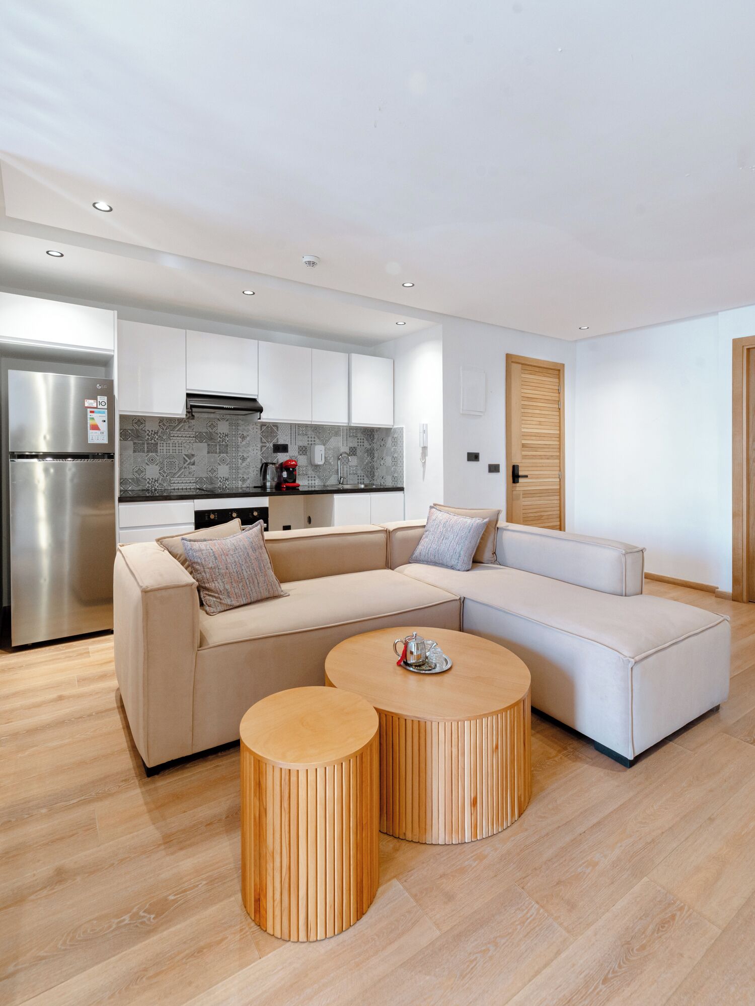 Una sala moderna con un sofá modular, dos mesas redondas de madera, una cocina con armarios blancos y un refrigerador de acero inoxidable.
