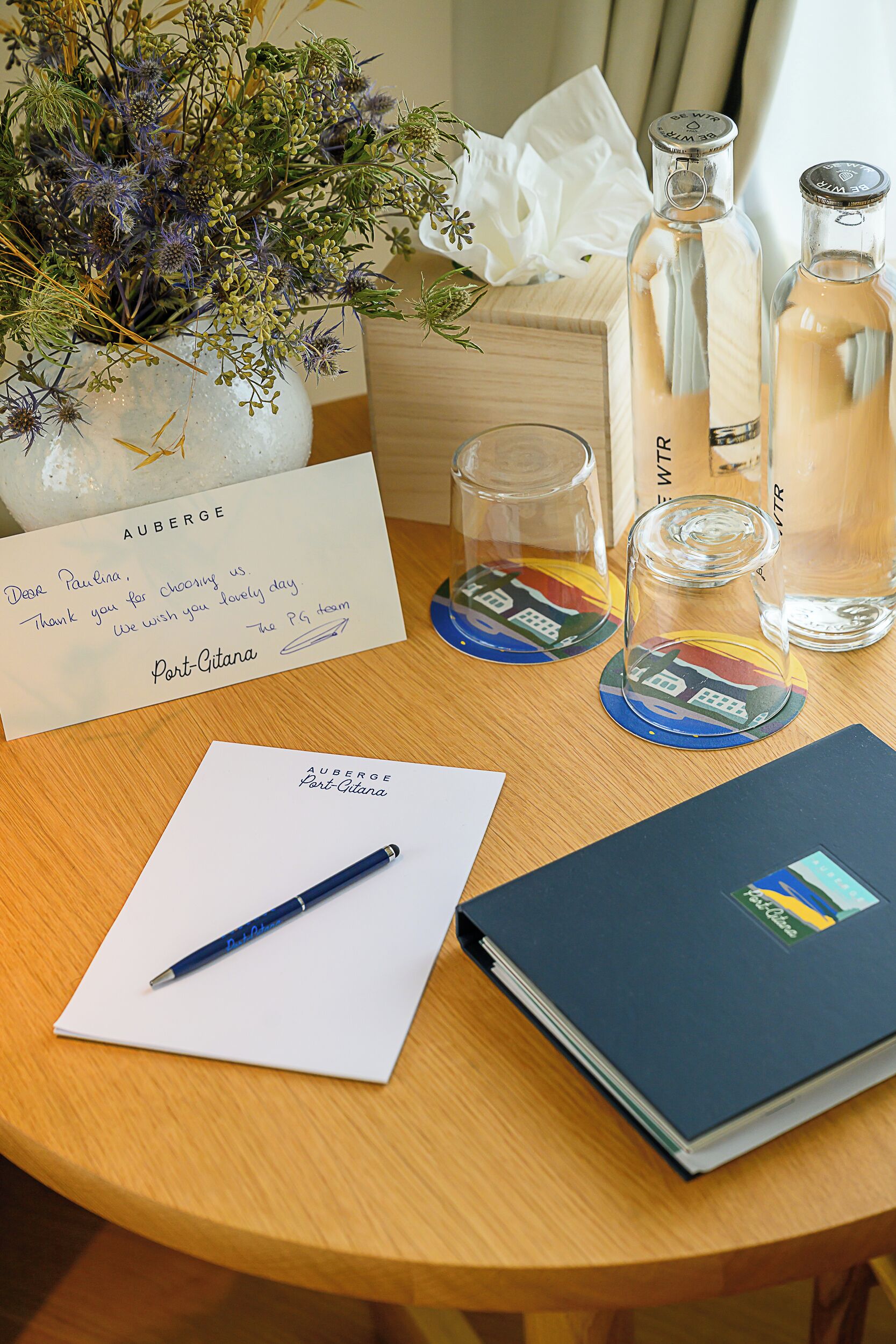 A wooden table with a vase of flowers, two bottles of water, a glass, a pen, a notepad, and a book.