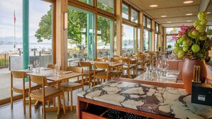 A restaurant with large windows, wooden tables, and chairs, overlooking a body of water. - Auberge Port Gitana (Bellevue)