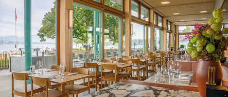 Een restaurant met grote ramen, houten tafels en stoelen, met uitzicht op een waterpartij.