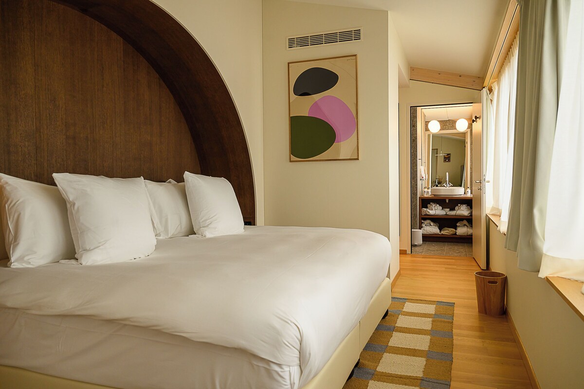 A neatly made bed with white linens, a wooden headboard, and a checkered rug on a wooden floor.