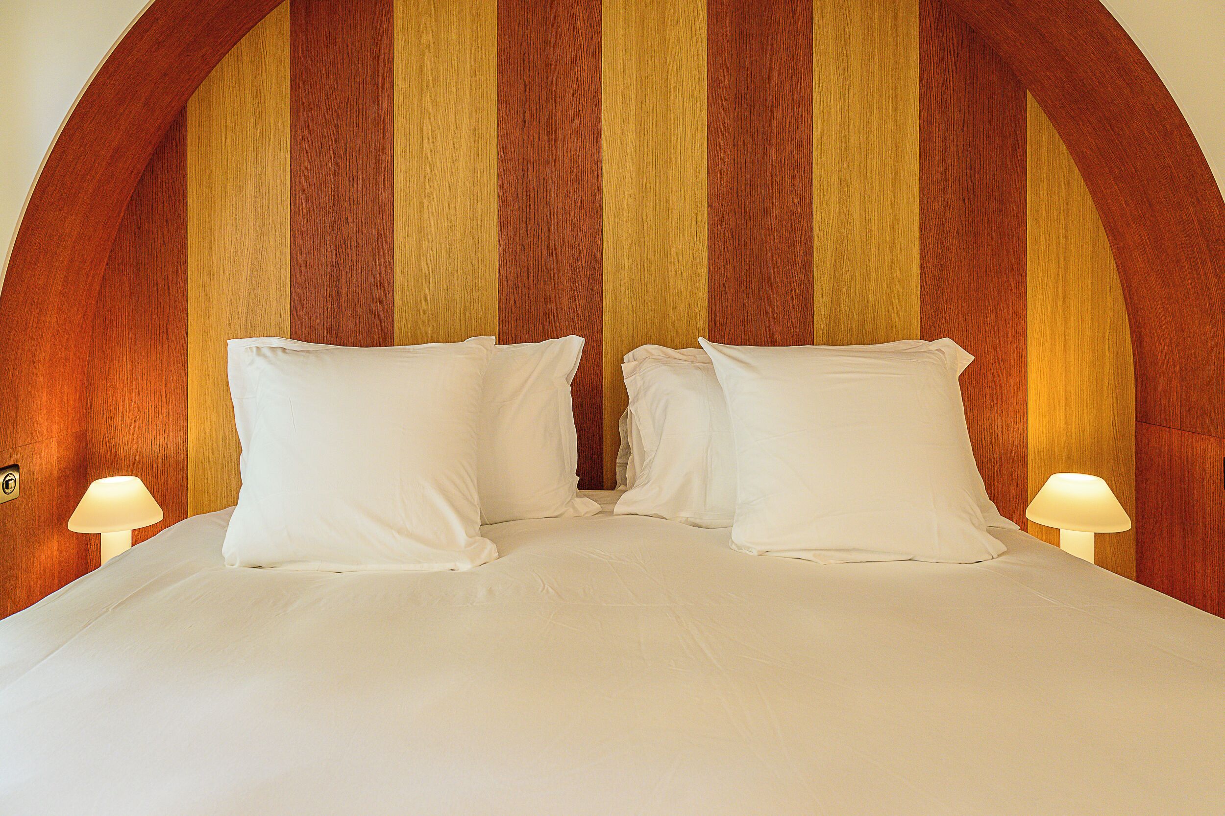 A bed with white linens and pillows, flanked by two bedside lamps, against a wooden headboard with vertical planks.