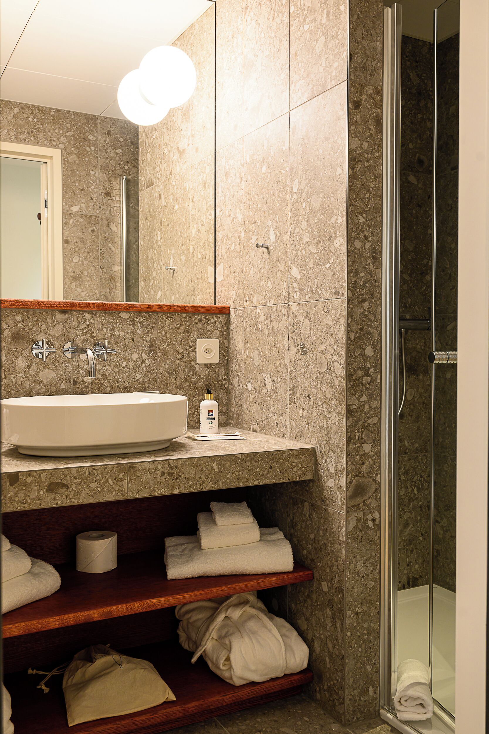 Ein Badezimmer mit einem runden Waschbecken, einem Spiegel, einer Dusche und Regalen mit Handtüchern.