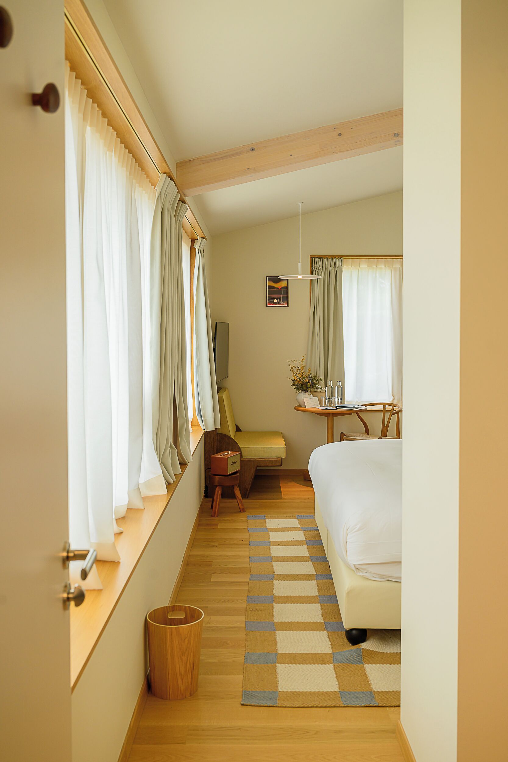 Una habitación de hotel con cama, un pequeño escritorio con silla, una ventana con cortinas, suelo de madera y una alfombra a cuadros.