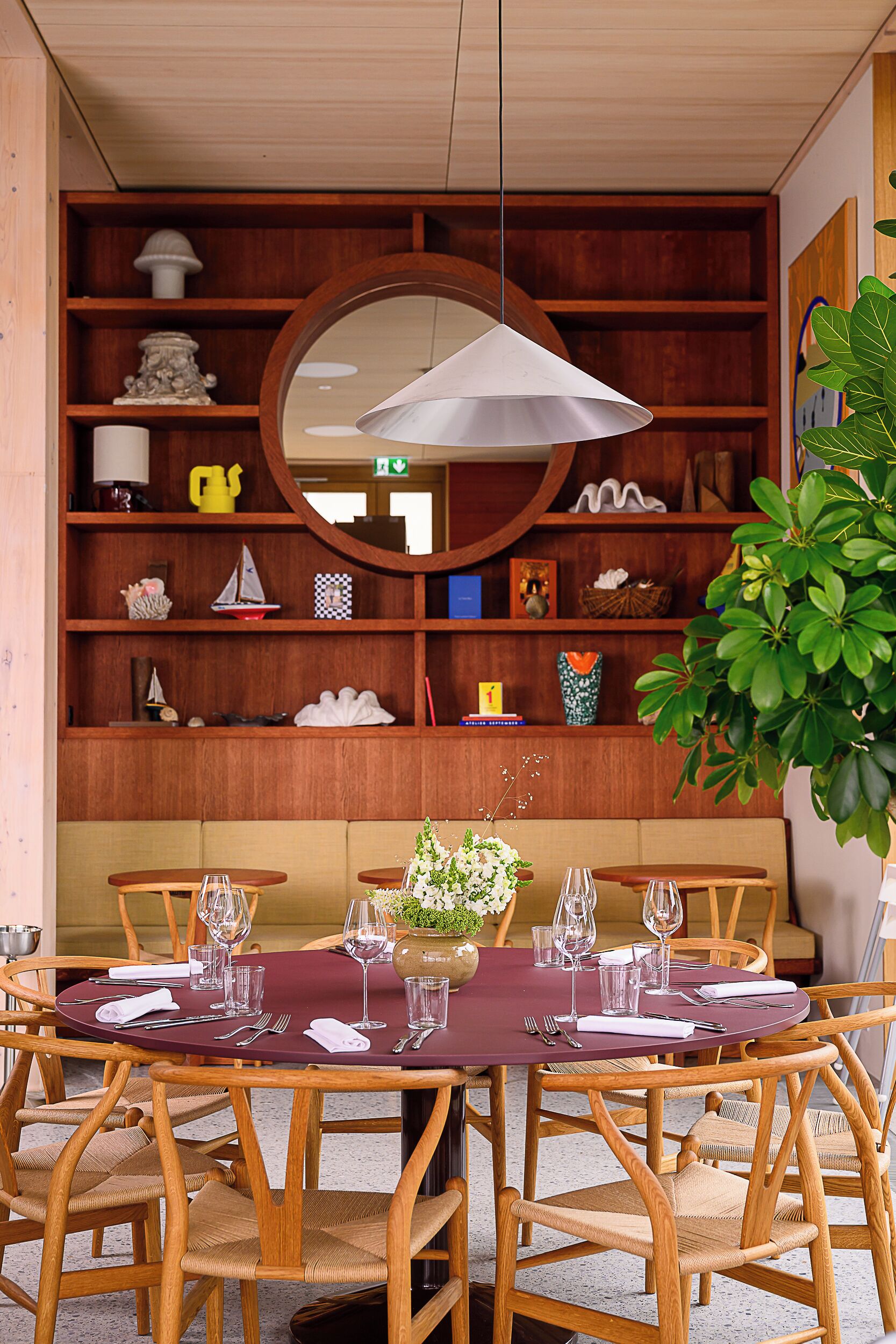 A dining area with a round table set for six, surrounded by wooden chairs with cushioned seats. A large mirror hangs above the table, and a potted plant is visible to the right.