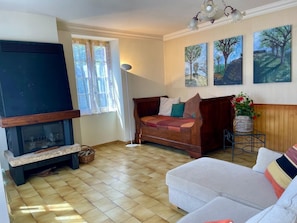 Un salon avec une cheminée, un canapé, une causeuse et un téléviseur. Il y a trois tableaux accrochés au mur.