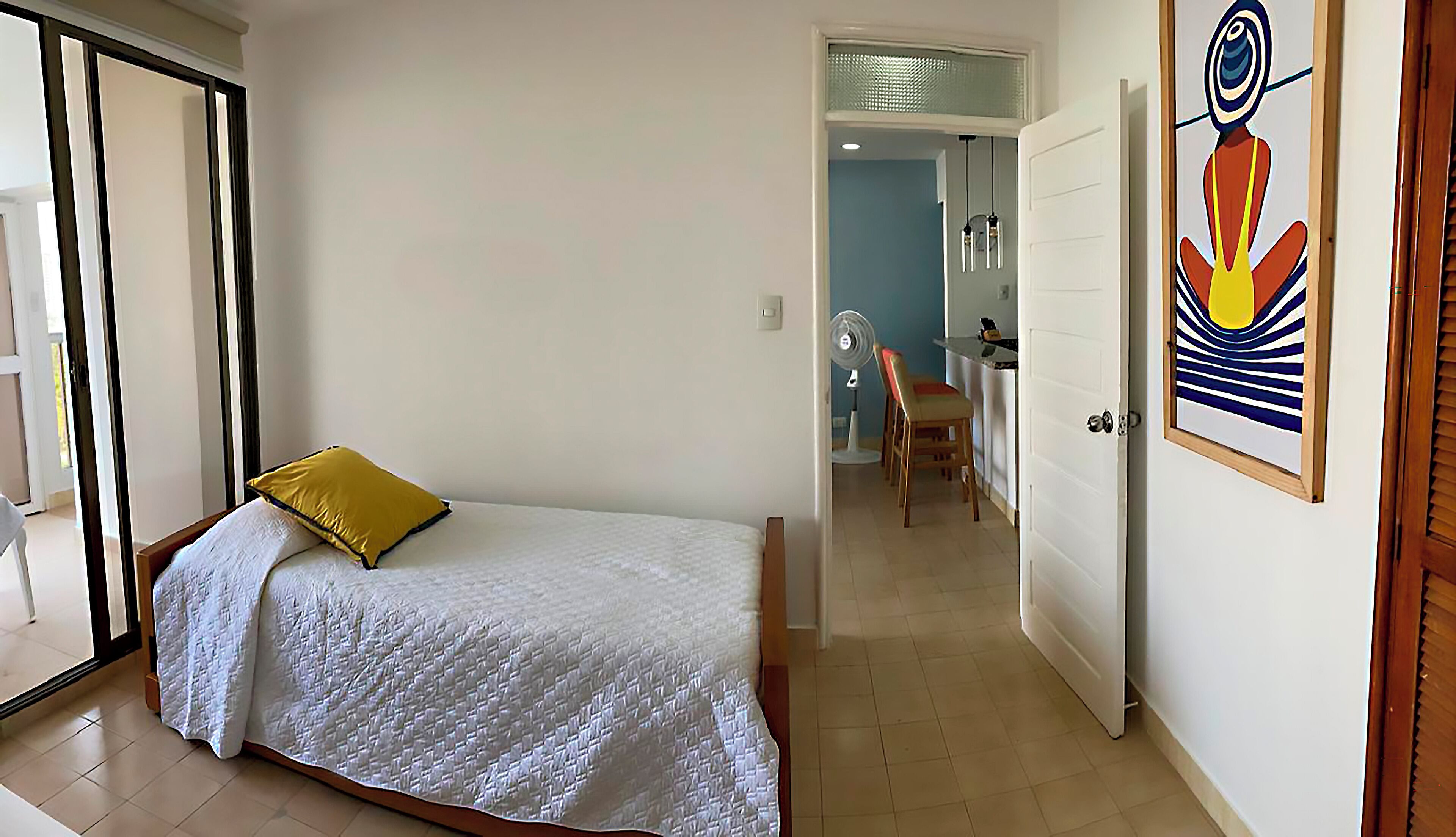 Ein Schlafzimmer mit Bett, einem gelben Kopfkissen, einer weißen Decke, einer Holztür und einem kleinen Tisch mit Stühlen.