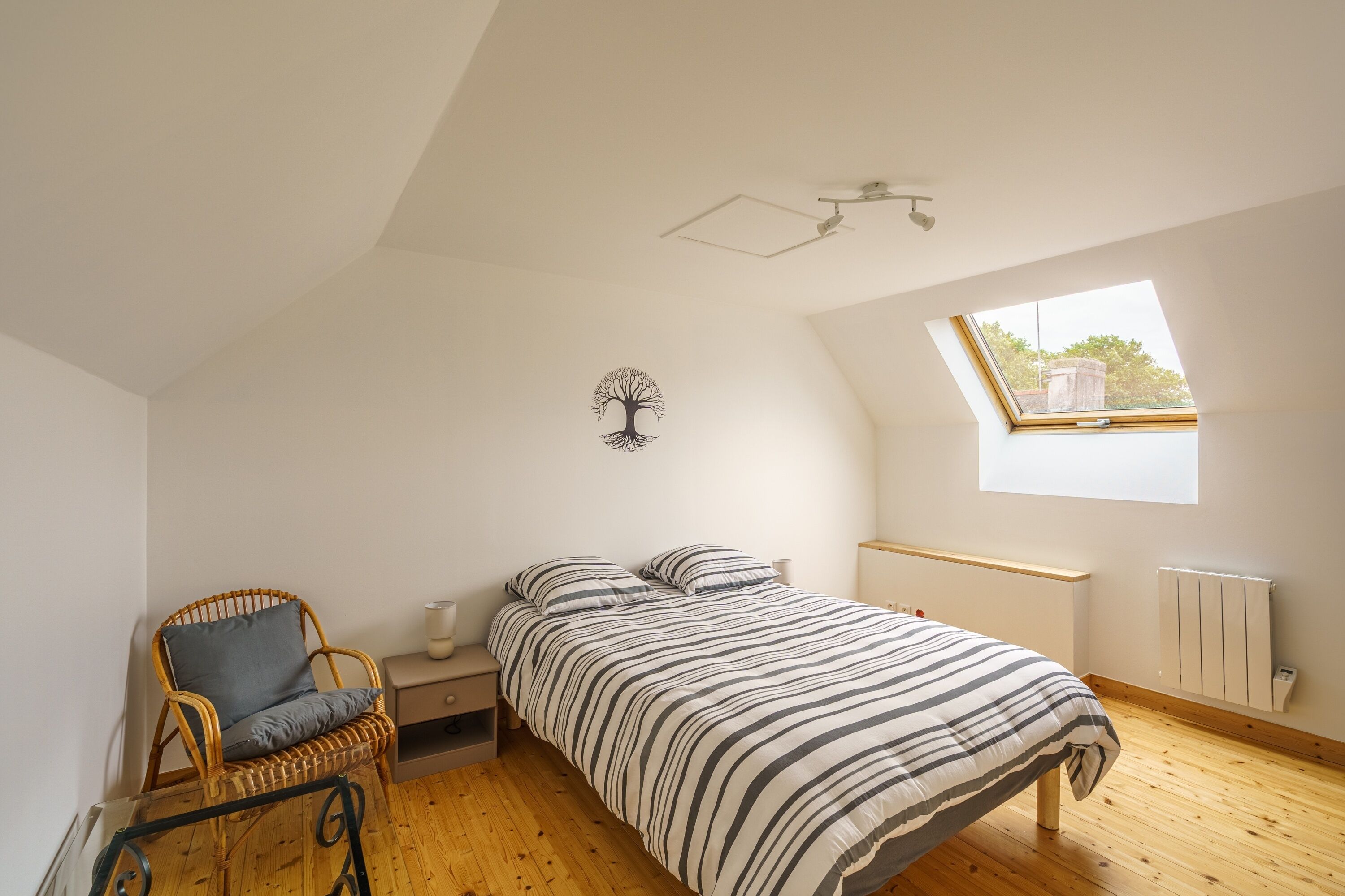 Ein Schlafzimmer mit schrägem Dach, einem Bett mit gestreifter Bettwäsche, einem Nachttisch, einem Sessel mit Kissen, einer Wanddekoration mit Baummotiv und einem Fenster mit Blick.