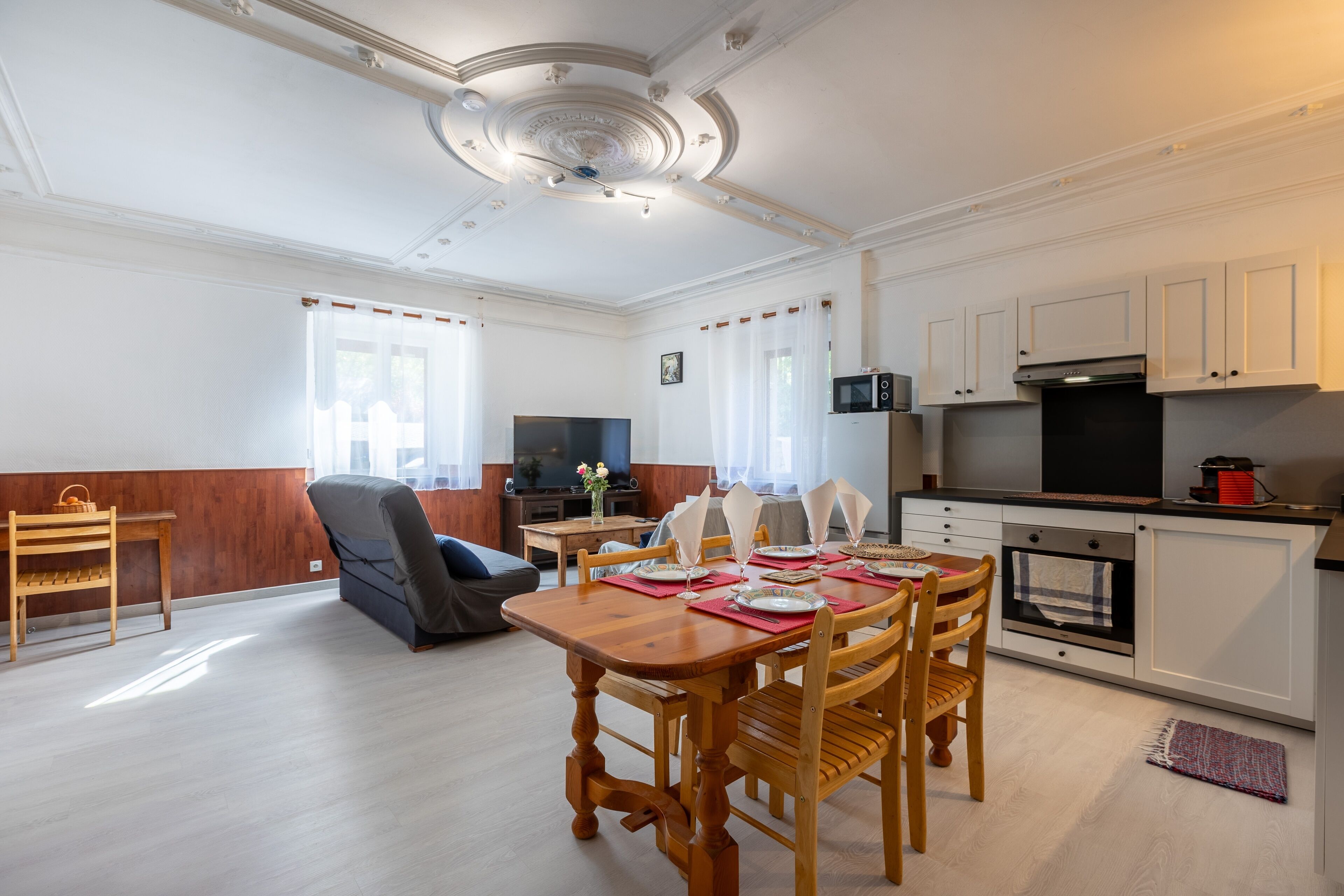 Une cuisine moderne avec des meubles blancs, un four à micro-ondes et un four traditionnel. On y trouve un coin repas avec une table et des chaises en bois, et un téléviseur en arrière-plan.