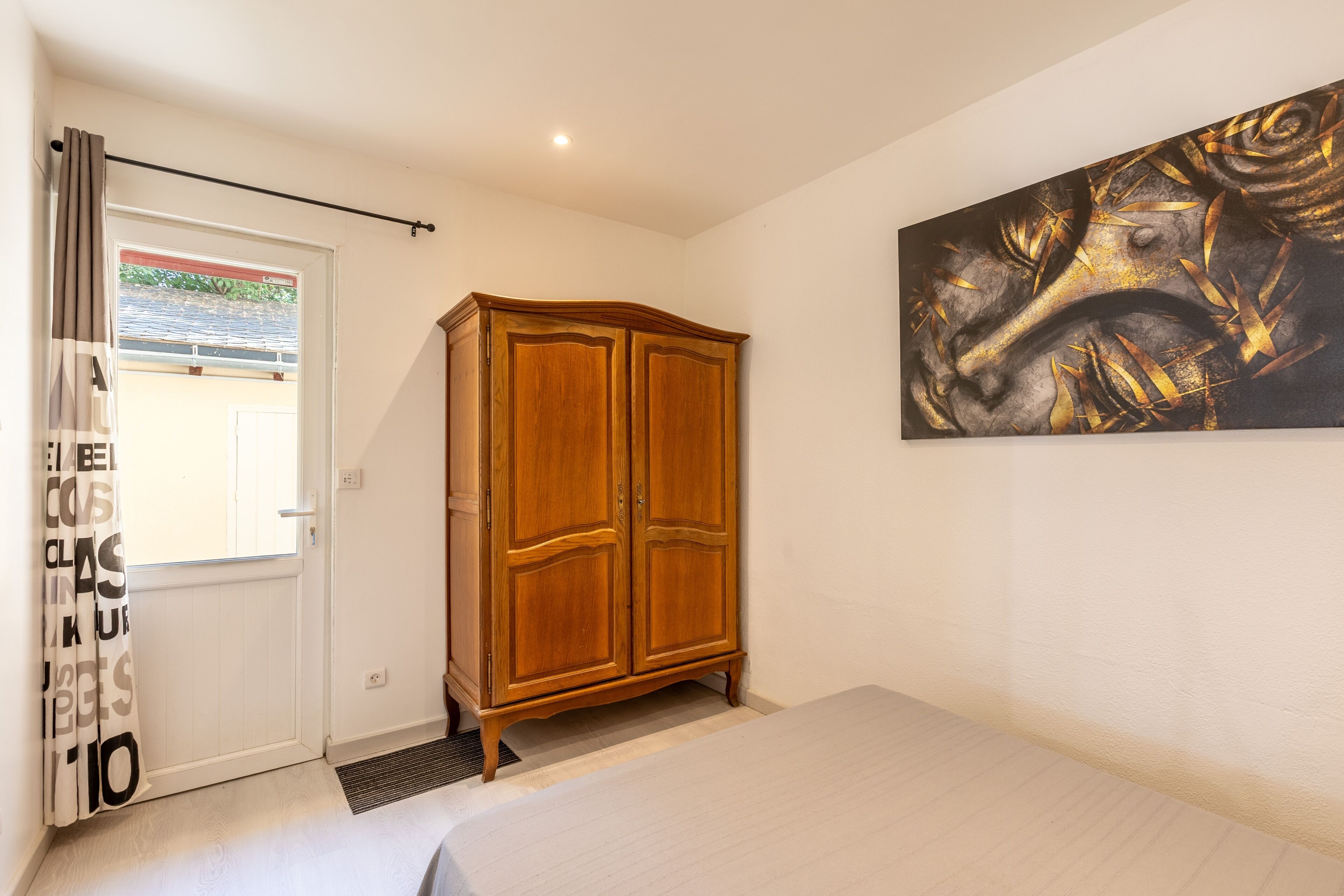 Um quarto com guarda-roupa de madeira, uma cama, uma janela com cortinas e um quadro pendurado na parede.