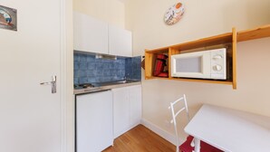 Eine kleine Küche mit weißen Schränken, einer blauen Spritzschutzleiste, einer Mikrowelle auf einem Holzregal und einem weißen Stuhl.