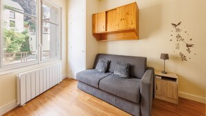 Ein Zimmer mit grauem Sofa, einem Holzschrank, einem kleinen Nachttisch mit Lampe sowie einem Fenster mit Blick auf Gebäude und Grünflächen.