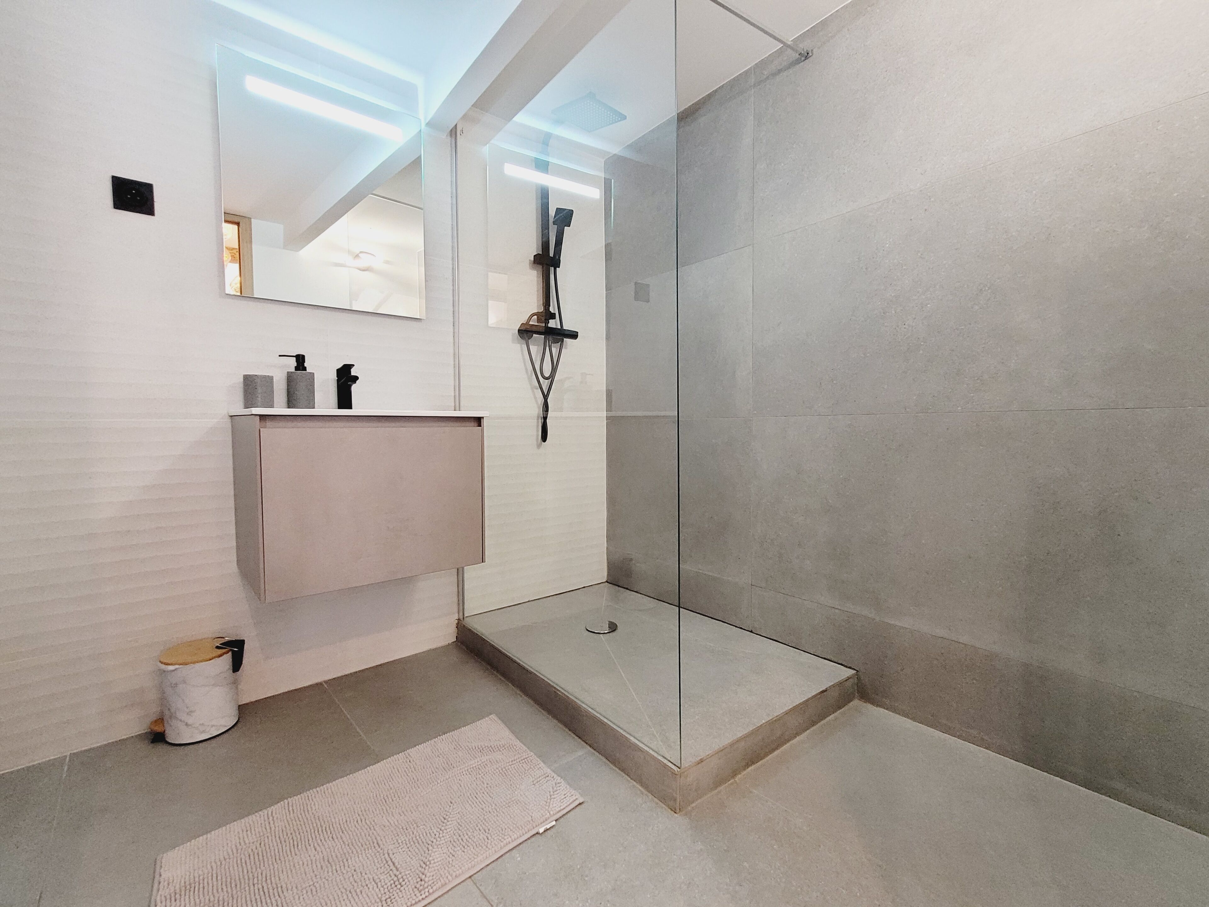 A modern bathroom with a glass shower enclosure, a sink with a mirror, and a small trash bin.