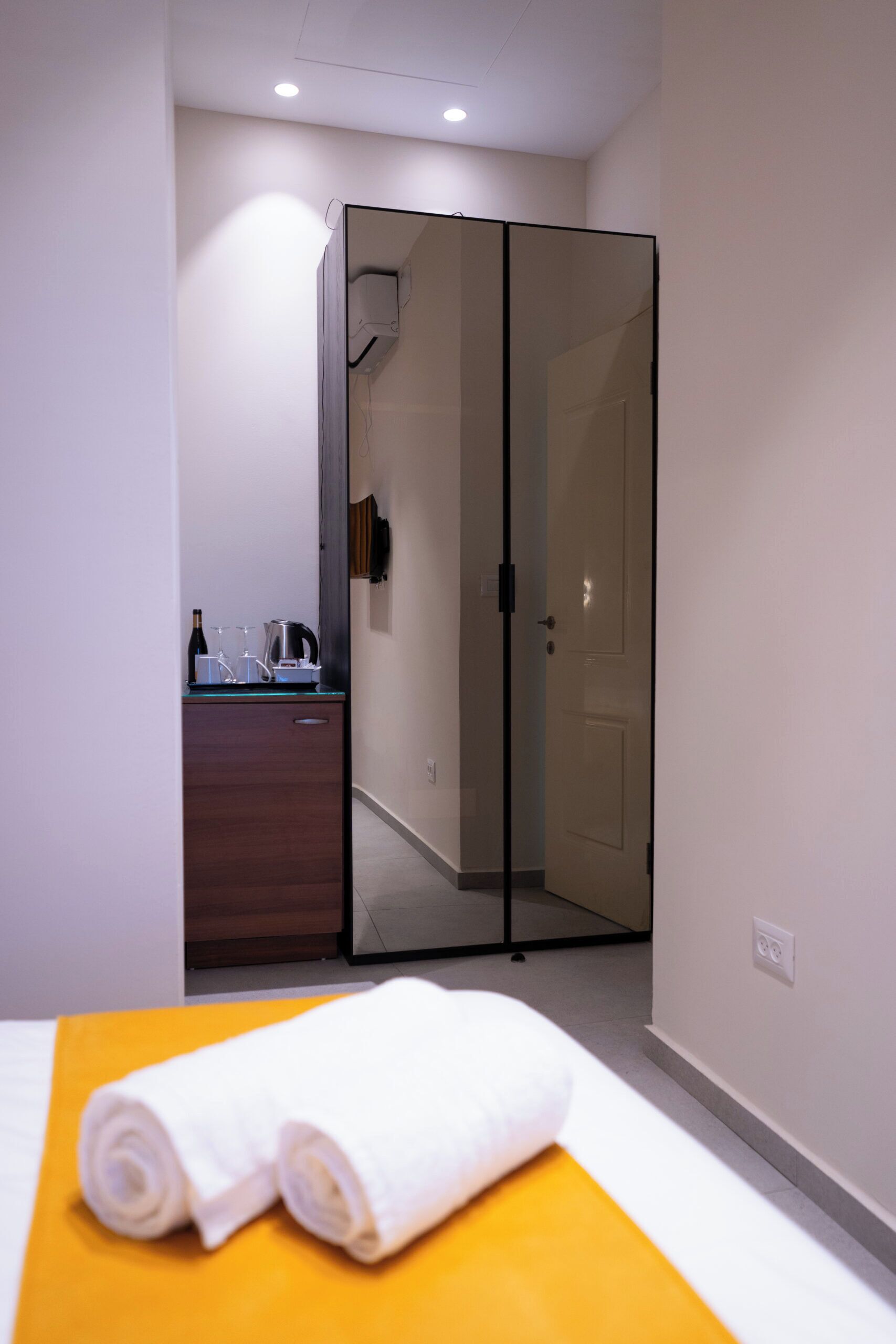 A hotel room with a bed, a yellow pillow, a white towel, a wooden dresser, a glass partition, and a door.