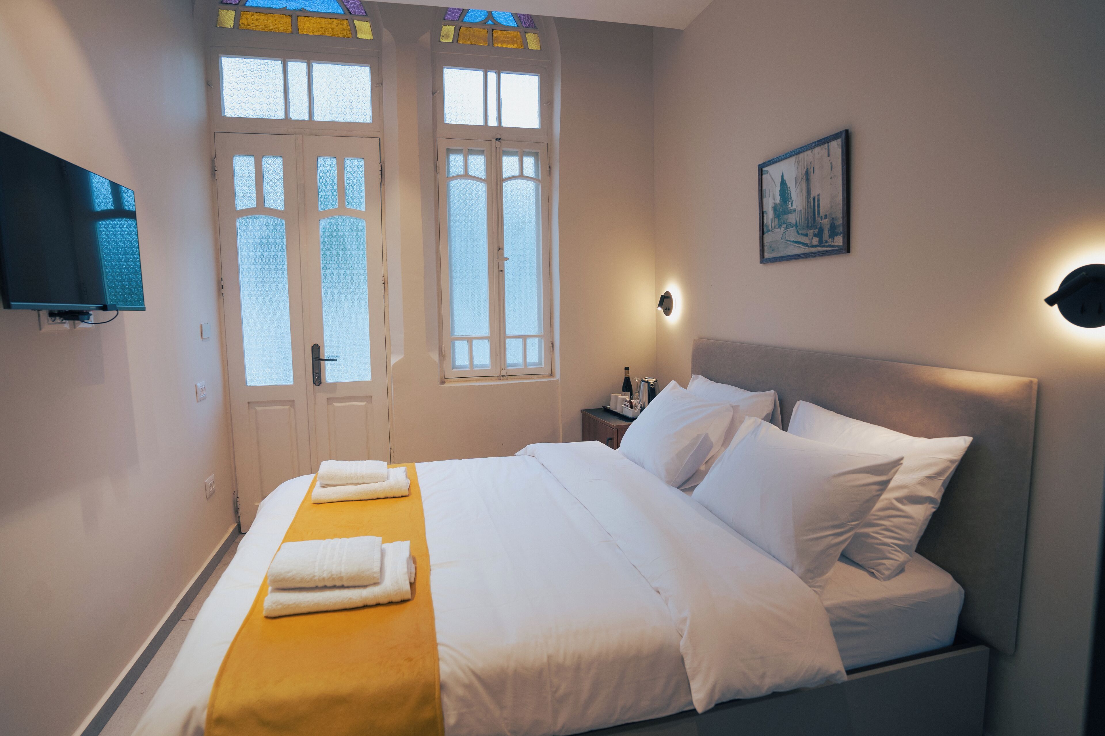 Una cama bien hecha con sábanas blancas y un detalle amarillo, una mesita de noche con lámpara y un cuadro en la pared.