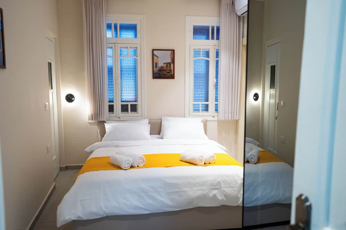 A hotel room with a large bed, two windows with curtains, a bedside table, and a mirror.
