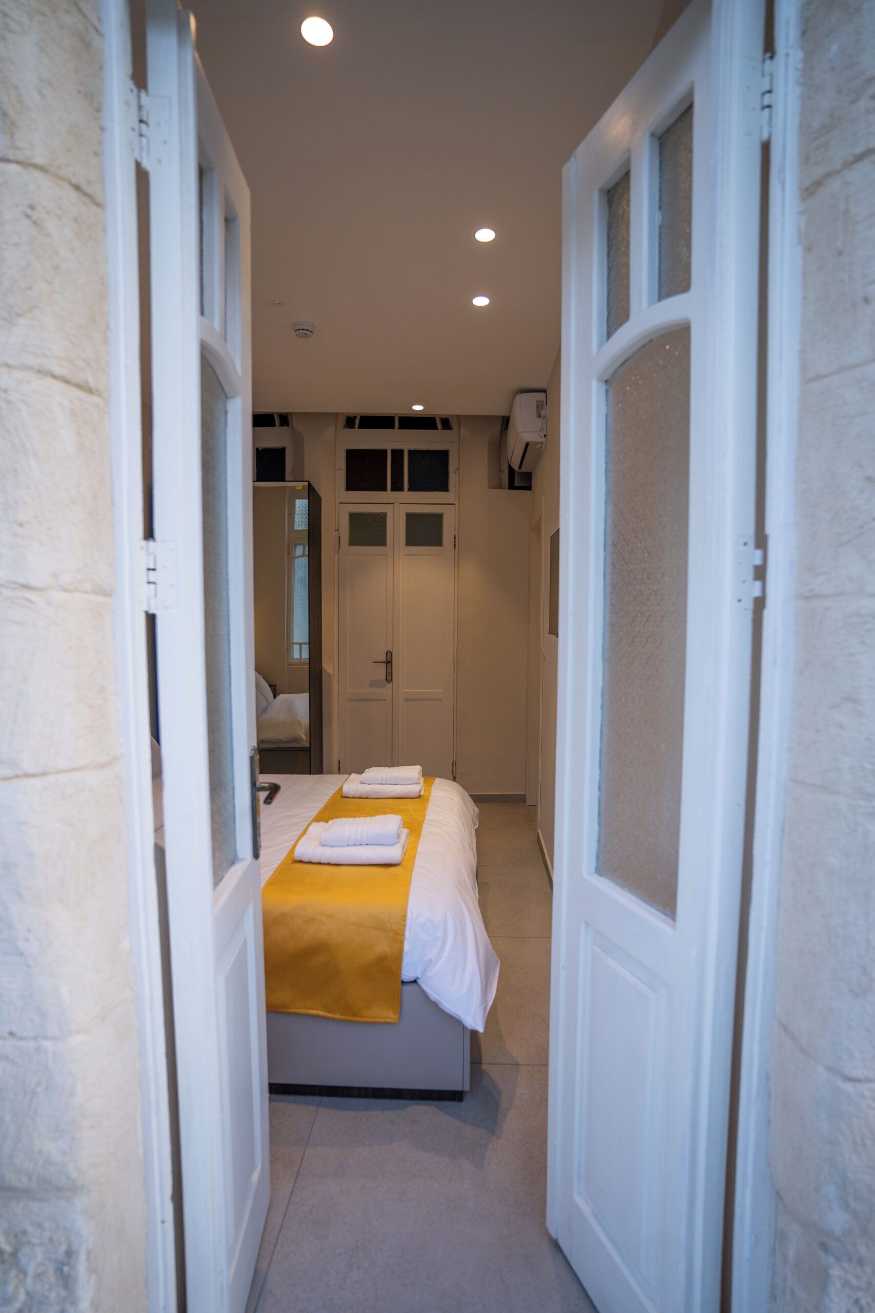 Un couloir étroit mène à une chambre à coucher où se trouvent un lit, une couverture jaune et une porte blanche.