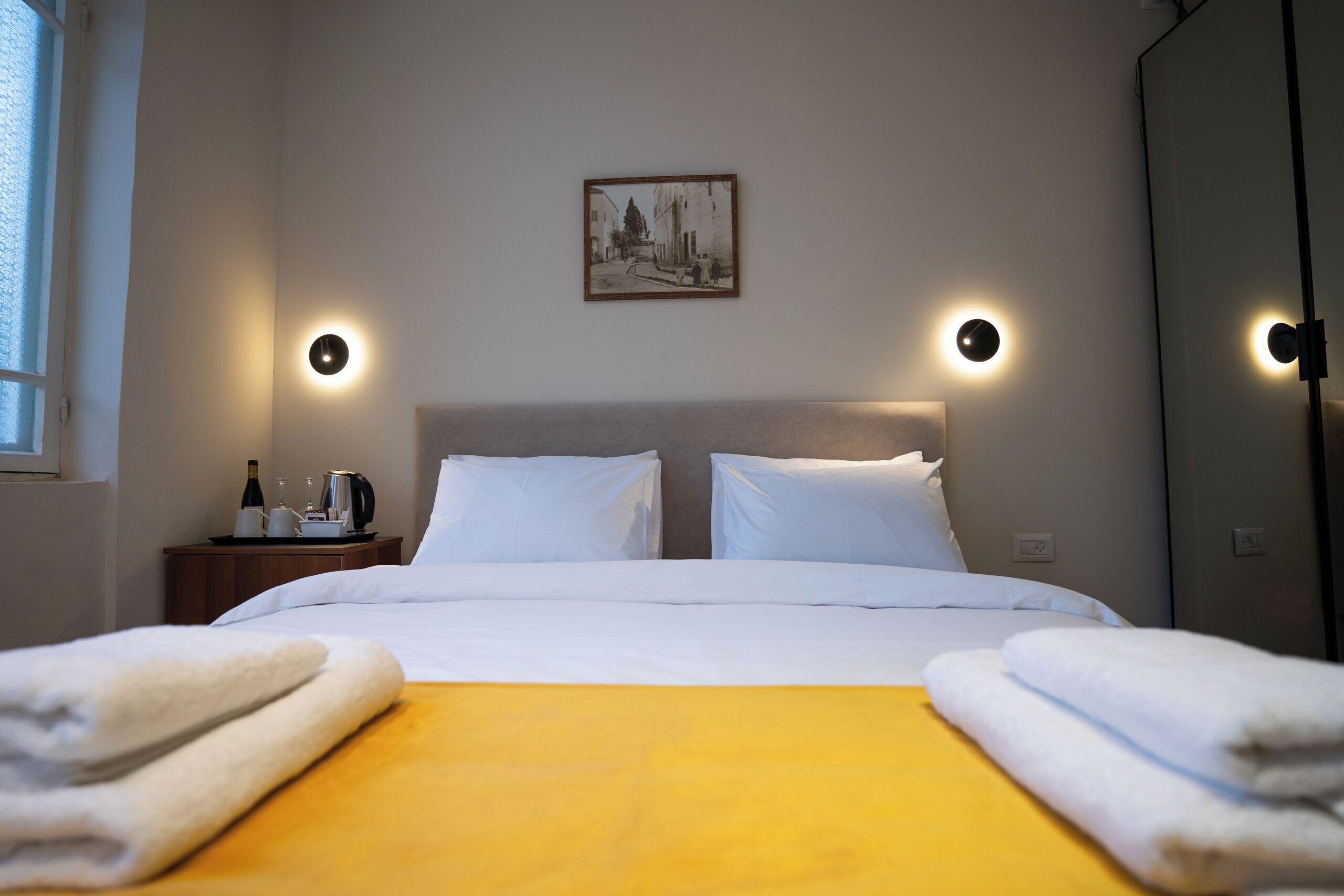Quarto de hotel com uma cama, almofadas brancas, um cobre-leito amarelo, uma mesa de cabeceira com uma garrafa e copos, e um quadro na parede.