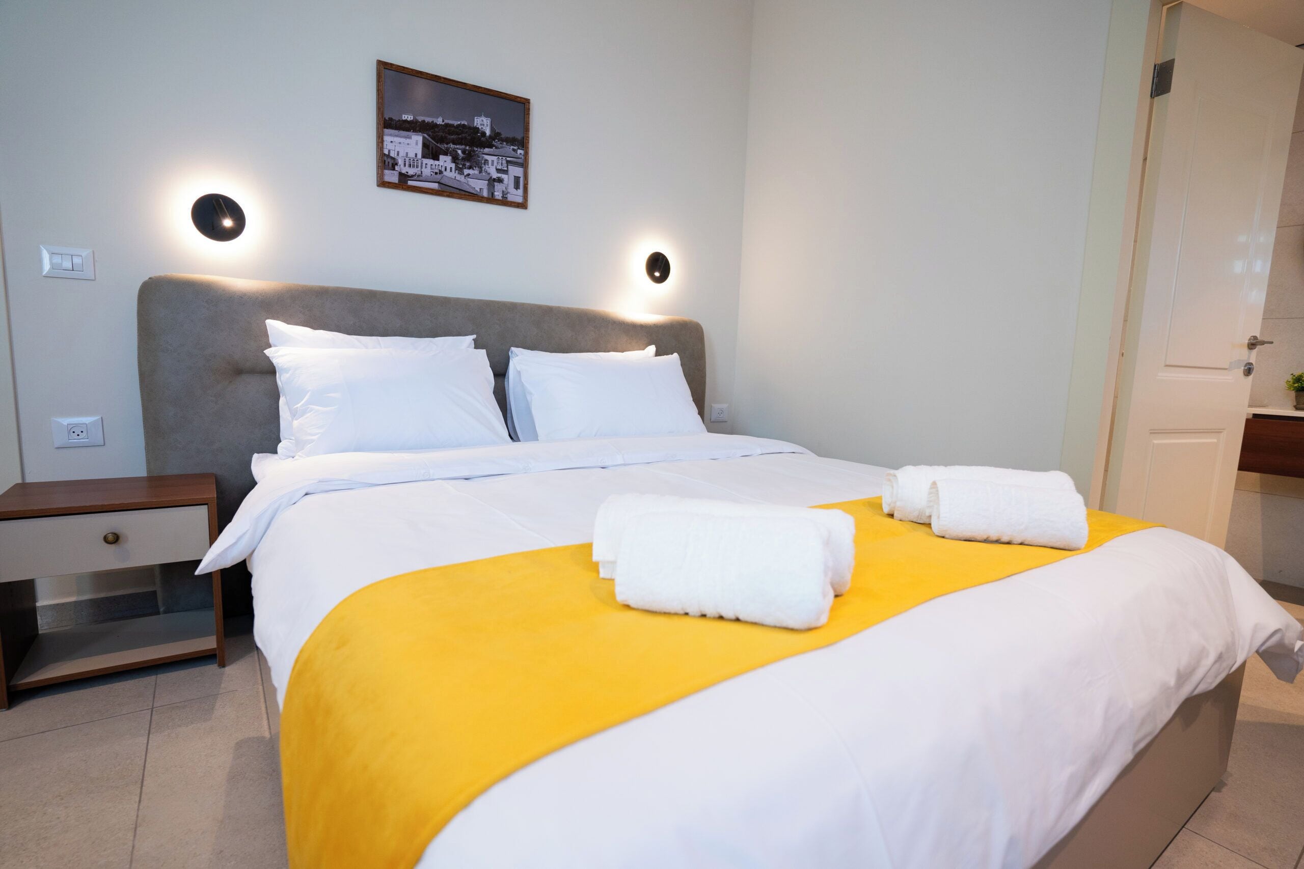 Un lit bien fait, avec du linge de lit blanc et un élément de couleur jaune, deux oreillers blancs et deux serviettes enroulées posées sur le lit.