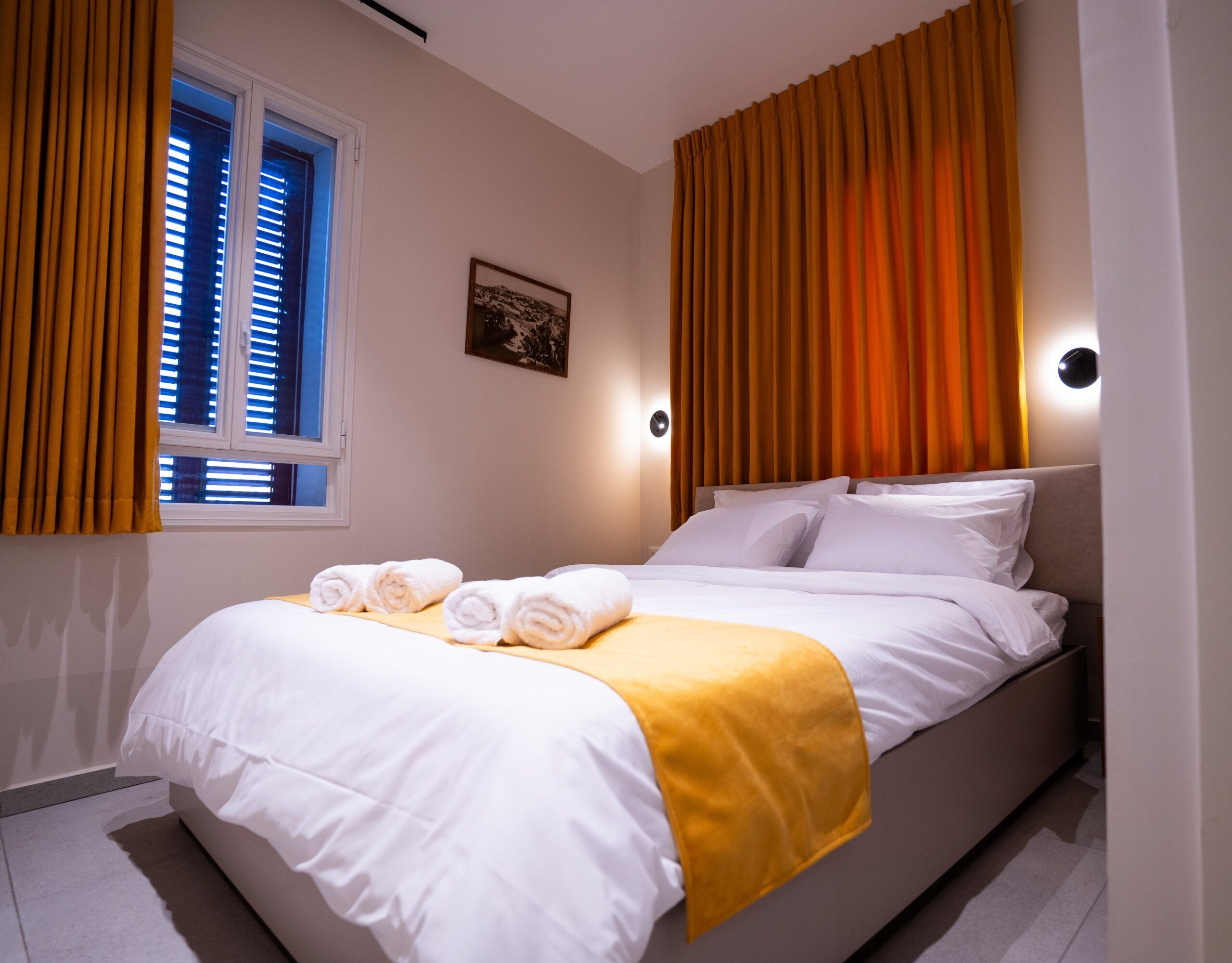 A neatly made bed with white linens and a yellow throw, a window with blinds, and a wall-mounted light.
