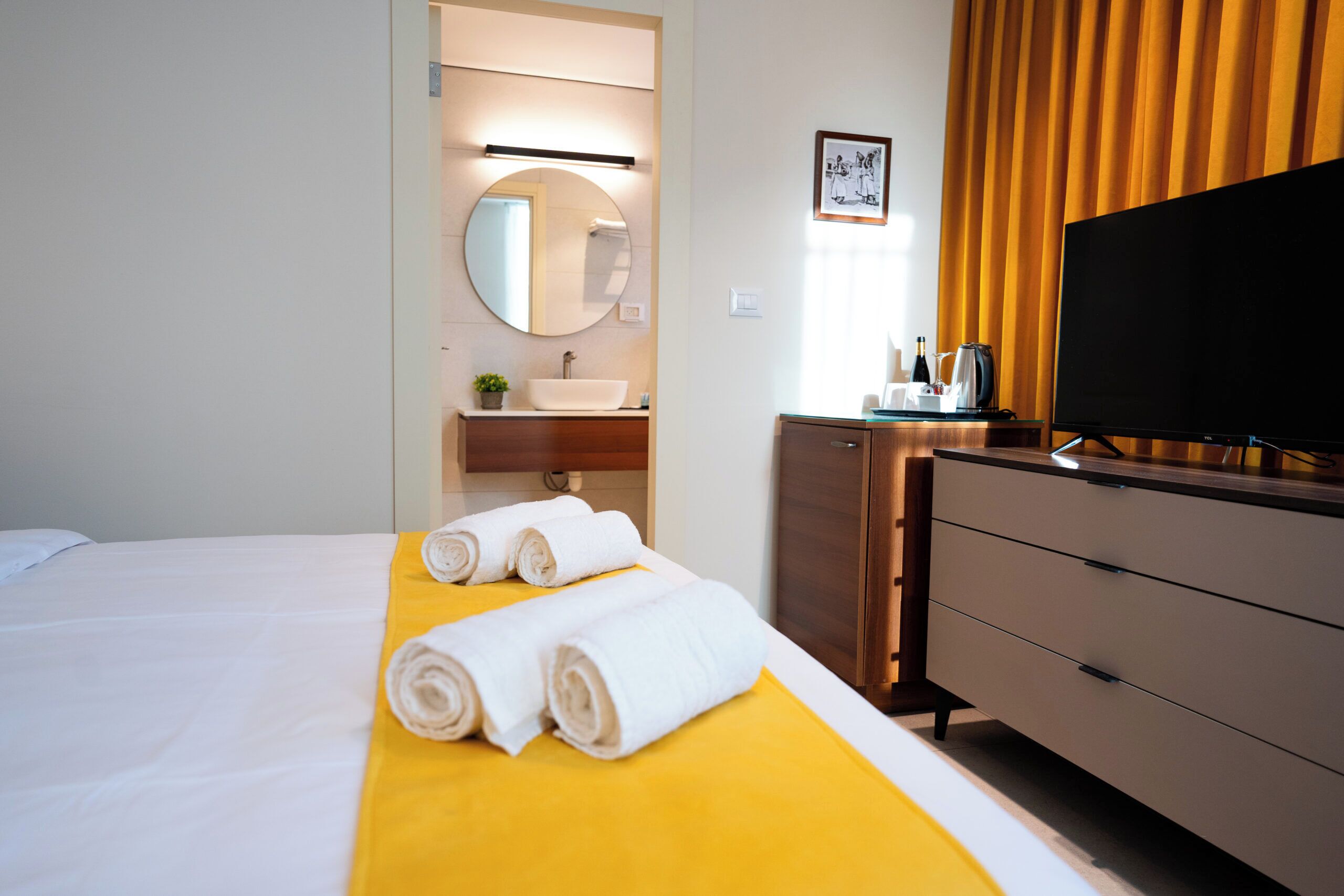 Une chambre d’hôtel avec un lit, une couvre-lit jaune, une télévision, une commode et une salle de bain avec un lavabo et un miroir.